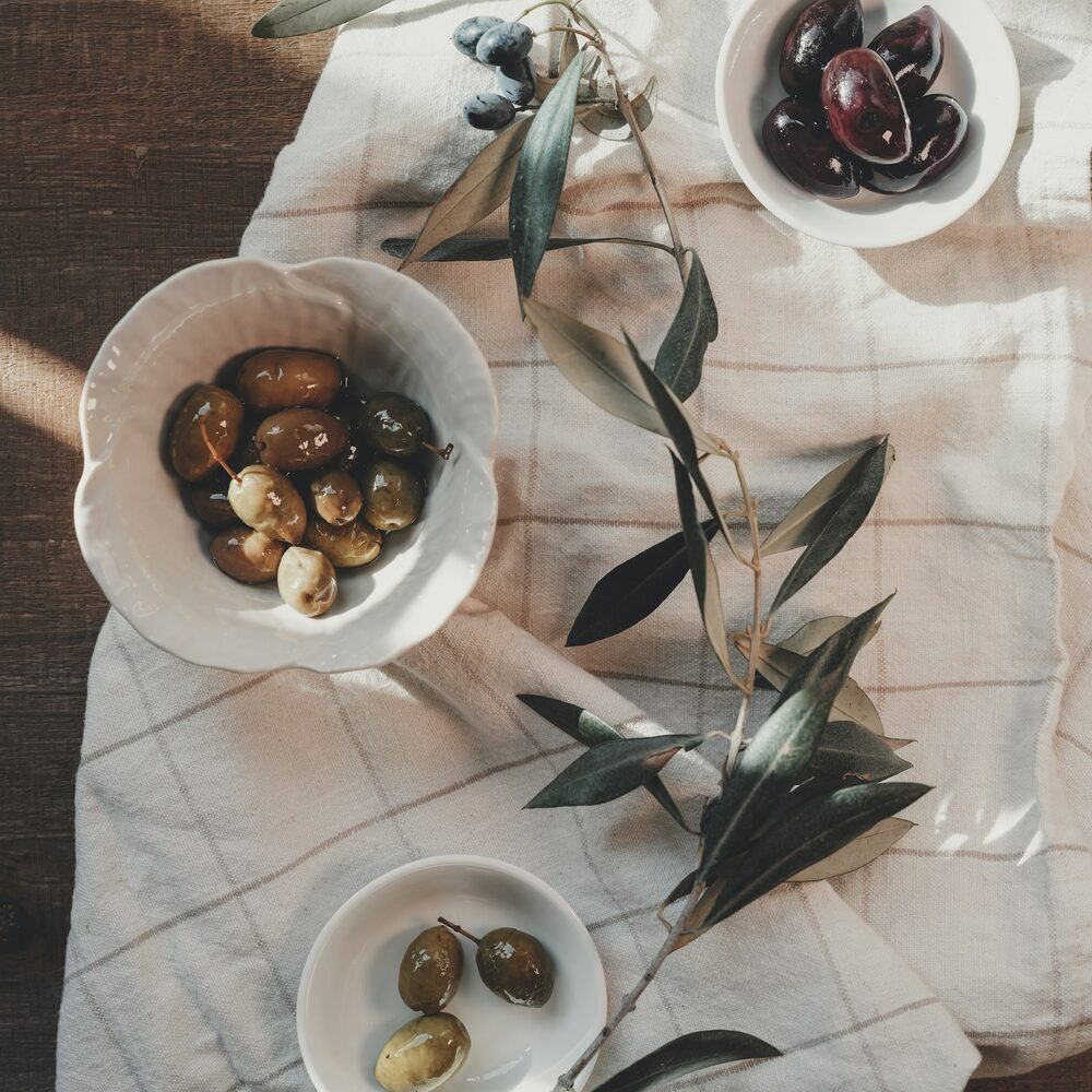 Trois coupelles d’olives violettes et brunes sur un torchon, rameau d’olivier