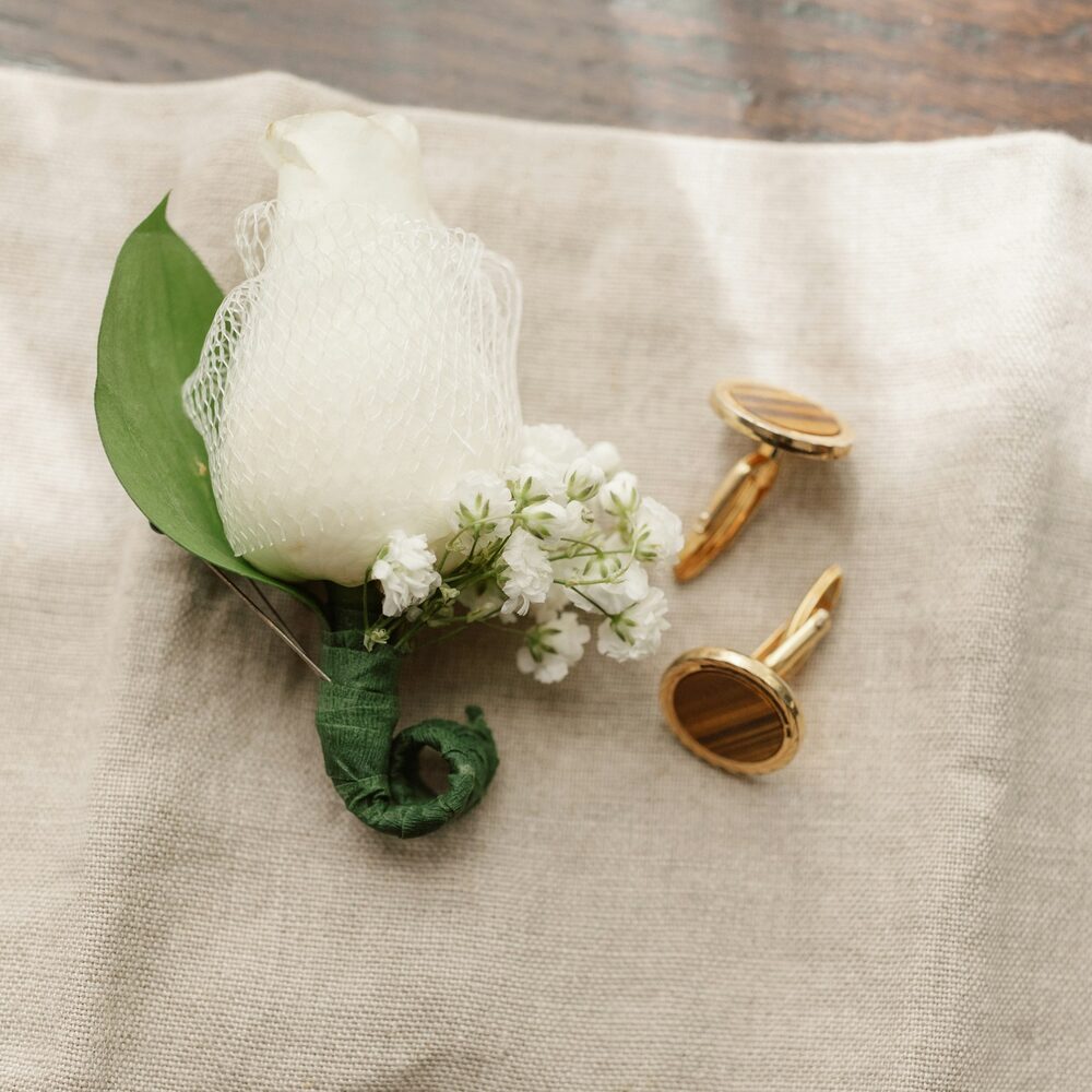 Boutonnière de rose blanche et de gypsophile posée sur du lin, à côté de boutons de manchette