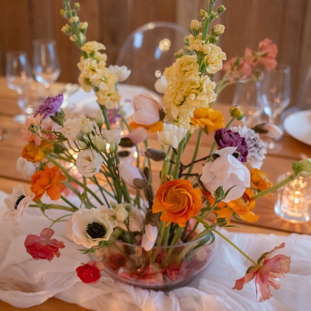 Centre de table de renoncules, giroflées et anémones dans une coupe en verre sur une table dressée