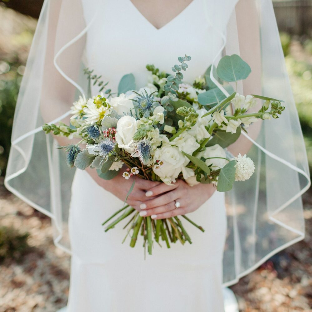 Mariée voilée tenant un bouquet blanc avec eucalyptus et chardons bleus