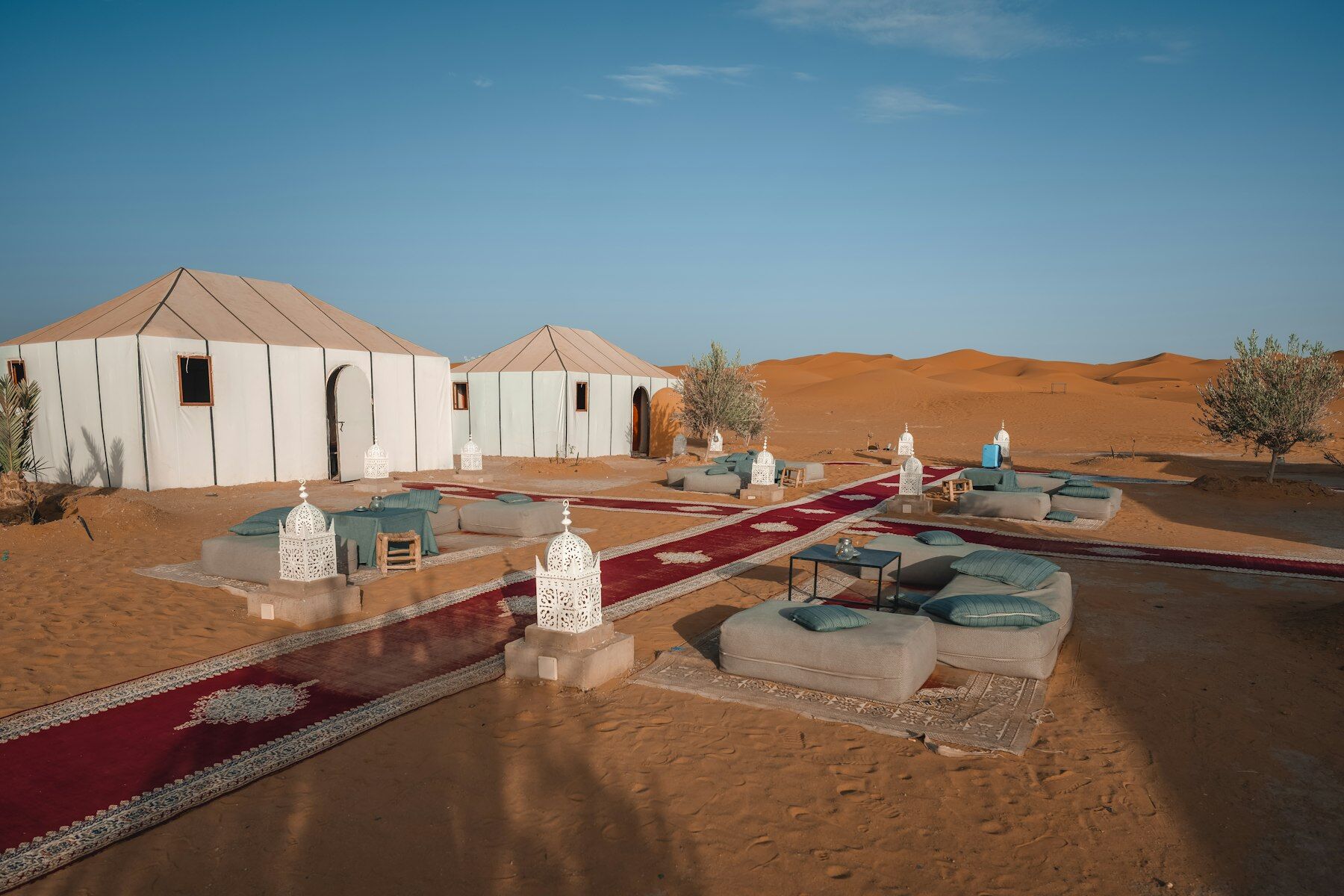 Tentes blanches d'un bivouac, tapis rouges et lanternes posés sur le sable, dunes au fond