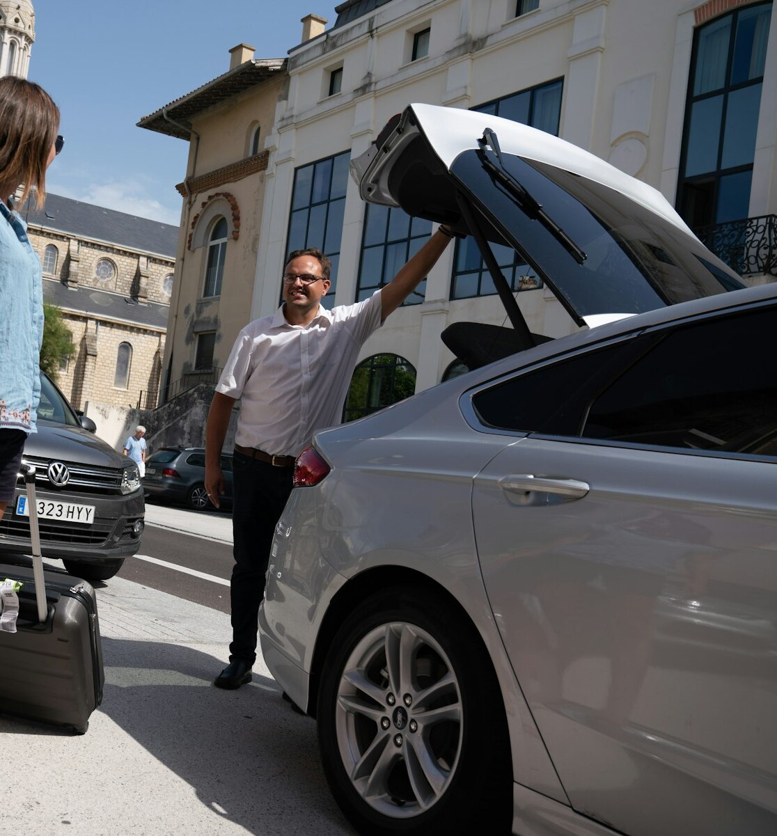 Un chauffeur ouvre le coffre d’une berline claire dans une rue